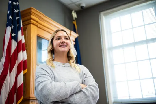A woman with long blonde hair stands confidently with her arms crossed, wearing a light gray sweater. She is positioned in a room with an American flag and sunlight streaming through the window blinds behind her.
