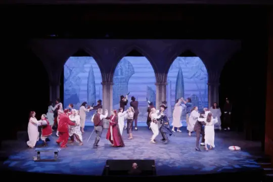 A theatrical performance with a large cast on stage, featuring actors dressed in medieval attire. The backdrop depicts Gothic arches and a cityscape. The scene is vibrant, showcasing the skill of Theatre Acting BFA students through dancing and lively interaction among the performers.