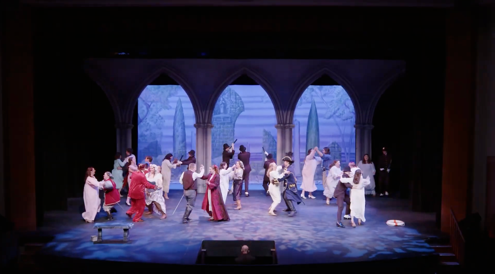 A theatrical performance with a large cast on stage, featuring actors dressed in medieval attire. The backdrop depicts Gothic arches and a cityscape. The scene is vibrant, showcasing the skill of Theatre Acting BFA students through dancing and lively interaction among the performers.