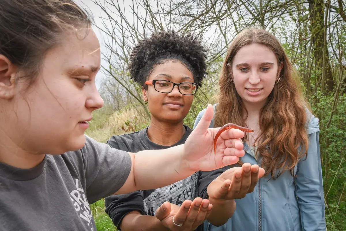 Three people stand outdoors, examining a small worm on one's hand. They are surrounded by greenery and trees. All are focused on the worm, showing a mix of curiosity and interest.