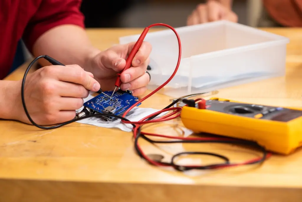 A person wearing a red shirt is using a multimeter with red and black probes to test a blue circuit board. The multimeter is yellow and sits on a wooden table, along with a clear plastic box.