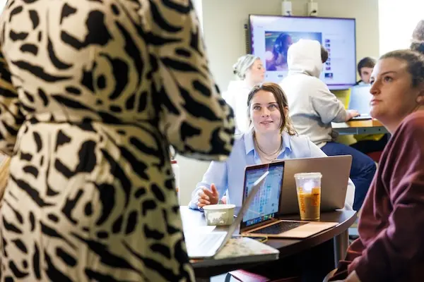 A group of people are collaborating at a round table with laptops and drinks. One person speaks, while others listen attentively. A large screen in the background displays images and text.