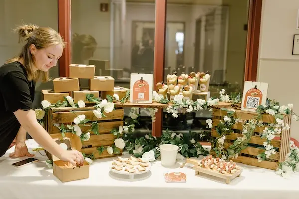 A woman arranges baked goods on a table decorated with wooden crates, white flowers, and greenery. Various pastries and boxed treats are displayed. A QR code and a sign with information are visible on the table.