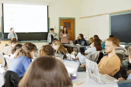 A classroom scene with students seated and engaged, some working on laptops. Three individuals stand at the front, one at a podium. Presentation slides and blackboards fill the background as part of a Political Science lecture or discussion.