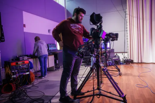 A man adeptly operates a professional video camera on a tripod in a studio with wood floors, crafting digital content. Another person tends to equipment amidst a tangle of cables and electronic devices. Light streams through the window, casting an inspiring glow on this hub of modern journalism.