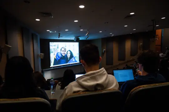 In a Film Studies lecture hall, students watch a video projected on the large screen. The film features two people bundled in blue jackets. Several students are seated with laptops open, engrossed in analyzing the scene attentively.