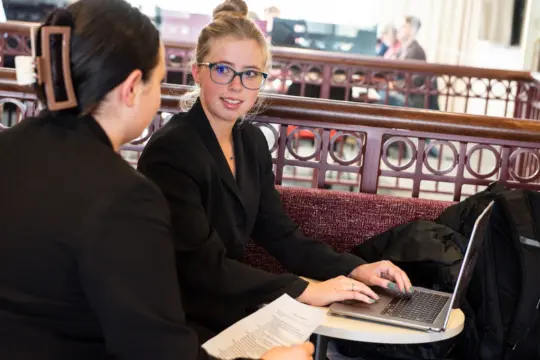 Two woman sit at a table in a cafe wearing suits. One is working on a laptop, wearing glasses and looking up, while the other holds a sheet of paper. They appear to be discussing something. The setting is casual with other people in the background.