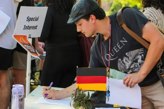 A person in a Queen T-shirt signs a paper at a table with a German flag. A "Special Interest" sign is visible nearby. People are around, and a small plant is on the table.