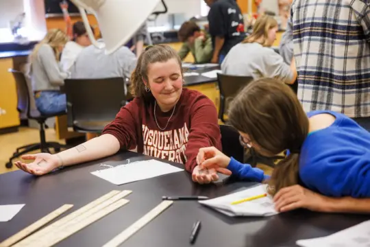 Students conducting a science experiment in a classroom. One student in a maroon sweatshirt holds out her arm while another takes a measurement. Other students in the background work together at desks.