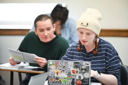 Two students sitting at desks in a classroom. One wears a cream beanie and a striped shirt, focused on a laptop covered with stickers. The other, in a green shirt, looks on while holding papers. A third student is blurred in the background.