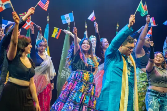 A diverse group of people on stage enthusiastically wave international flags. They are dressed in colorful, traditional costumes, creating a vibrant and festive atmosphere. The background shows a row of flags from various countries.