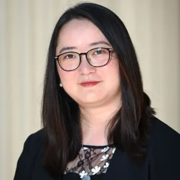 A person with long, dark hair and glasses is wearing a black top with a floral design at the neckline. The background is a light, neutral color.