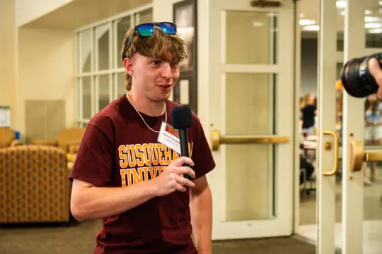 A young person with sunglasses on their head holds a microphone while speaking, wearing a maroon "Susquehanna University" t-shirt. They are indoors, with a blurred background showing couches and people.