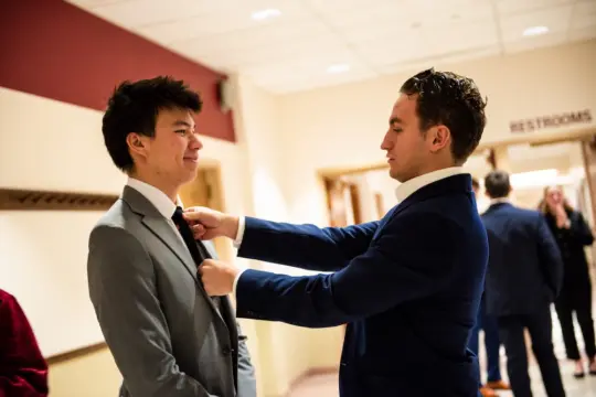 Two men in suits in a brightly lit hallway. One adjusts the tie of the other, who smiles. A "Restrooms" sign is visible on the wall, and there are blurred figures in the background.