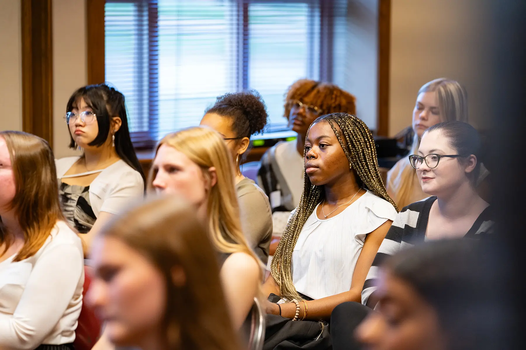 A diverse group of people attentively listening in a room with wooden blinds. They are seated closely, some with notebooks, indicating a learning or seminar setting. The lighting is warm, highlighting a focused and engaged audience.