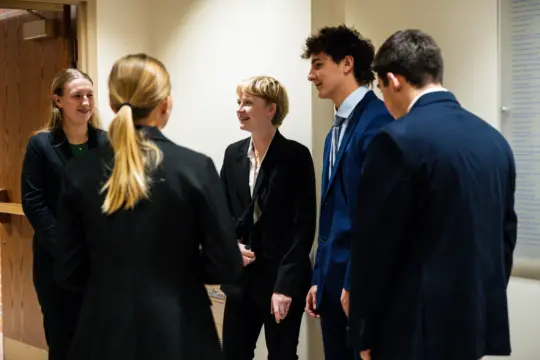 Five people in formal attire stand in a hallway, engaged in conversation and smiling. Three face the camera, while two have their backs to it. The setting appears to be inside an office or Legal Studies department at a school.
