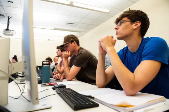 Students sit at desks with computers, focused on a mathematics presentation in a classroom setting. They have notebooks and water bottles; one wears a cap and glasses, another wears a blue shirt. The room has bright lighting and a projector on the ceiling.