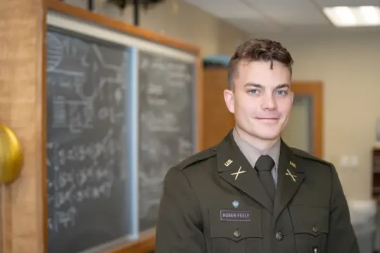 A person in a military-style uniform stands in a classroom in front of a chalkboard filled with writing and diagrams. The person is smiling slightly and looking at the camera. The classroom is well-lit with fluorescent lighting.