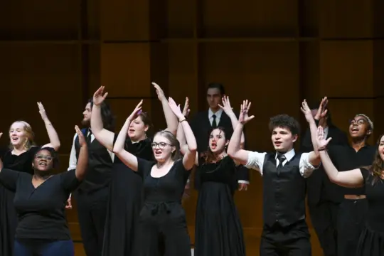 A group of performers, dressed in black attire, stand on stage with their arms raised in animated gestures. They appear to be engaged in a lively music performance, with one person in a suit seen in the background. The setting is a theater with a wooden backdrop.