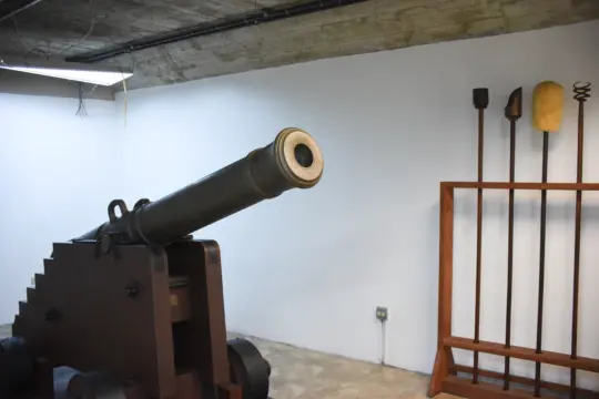 A historic cannon is displayed indoors, mounted on a wooden base with wheels. In the background, various tools or equipment are neatly arranged on a wooden rack against a white wall.