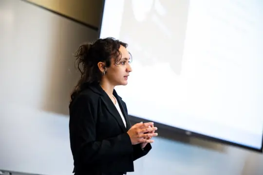 A woman in a black blazer is standing and speaking in front of a projection screen in a bright room. She appears to be giving a presentation or lecture.