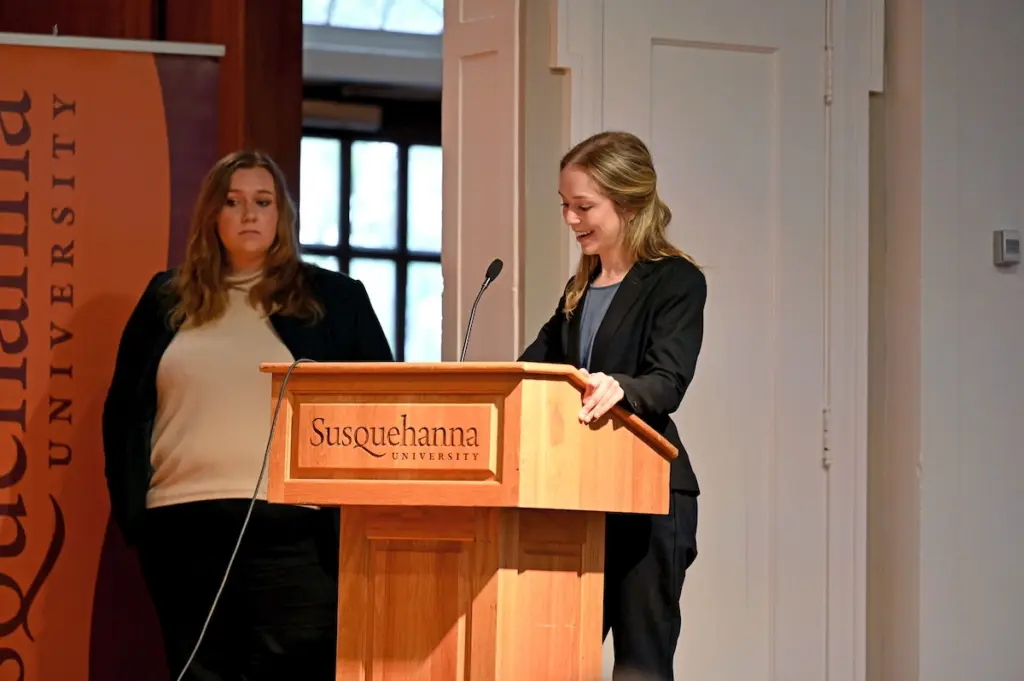 Two women at a podium with "Susquehanna University" on it. One is speaking into a microphone, wearing a dark suit. The other stands nearby, wearing a white shirt and dark jacket, with a Susquehanna University banner in the background.