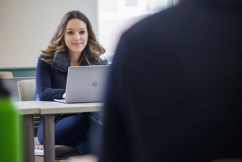 A woman with long hair is sitting at a desk using a laptop. She is smiling and facing another person who is partially visible in the foreground. The setting appears to be a classroom or office.