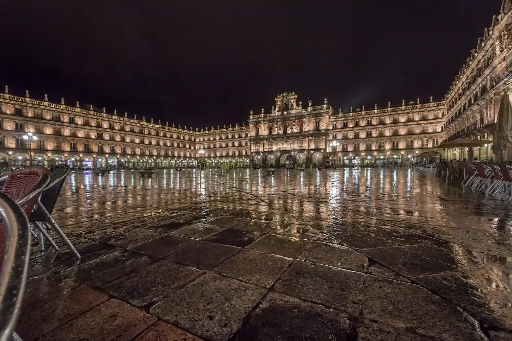 The image shows a wet, reflective Plaza Mayor at night, with ornate, illuminated buildings surrounding the square. Empty outdoor chairs are visible in the foreground, adding to the serene atmosphere.