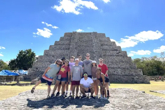 A group of people posing in front of a large stone pyramid under a clear blue sky. Some are wearing hats and sunglasses, and all are casually dressed. They stand on a stone platform with trees in the background.