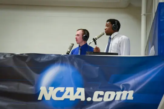 Two commentators wearing headphones and ties speak into microphones at a sports event. A large banner with "NCAA.com" is displayed in front of them. They are in a broadcast booth setting.
