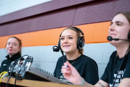 Three people are seated at a table with audio equipment. The person in the center is smiling and wearing a headset. The two others, also wearing headsets, are engaged in conversation. The background features a two-tone wall with maroon and orange stripes.