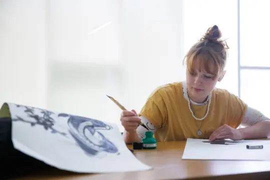A person with a bun hairstyle is seated at a wooden table, dipping a quill pen into green ink. They are focused on artwork in front of them, with a completed design featuring a large snake nearby. Soft, natural light fills the room.