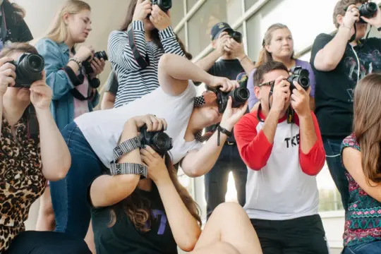 A group of people crouched and standing, all holding cameras, focus on something outside the frame. They are gathered in an outdoor space with a reflective glass building in the background.