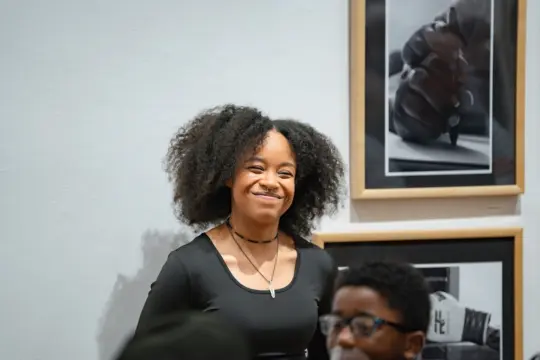 A person with curly hair smiles brightly while standing in front of two framed black-and-white photographs on a wall. The mood is positive and inviting.