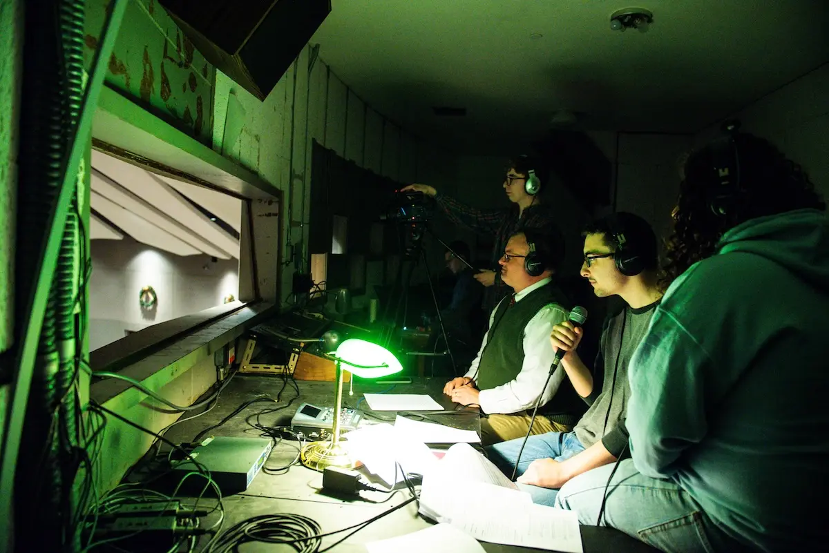 Four people wearing headsets are seated at a dimly lit control booth with a window overlooking a stage. One person holds a microphone, and papers are spread across the desk, illuminated by a green desk lamp.