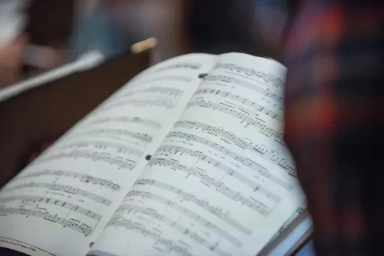 An open book displaying pages of sheet music, intertwined with the essence of collaborative piano, features musical notes and staff lines. The blurred background hints at a person in a plaid shirt, suggesting a rehearsal setting rich with harmonious synergy.