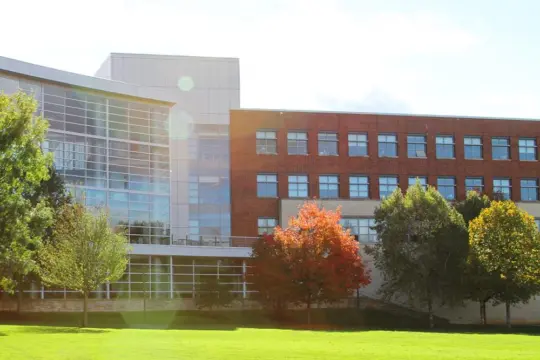 A modern building with glass and brick facade is surrounded by lush green grass and trees with fall foliage. Sunlight creates a lens flare effect in the foreground.