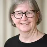 A smiling woman with short gray hair and glasses is wearing a black top. She stands against a light-colored background.