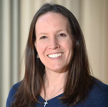 A woman with long brown hair is smiling at the camera. She is wearing a blue top and a cross necklace. The background is softly blurred.