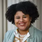 A person with curly hair smiles, wearing a light green blazer, patterned shirt, and pearl necklace. The background features light-colored vertical blinds.