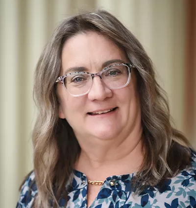A person with long wavy hair and glasses smiles at the camera, wearing a floral patterned blouse and a gold necklace. The background is softly blurred.