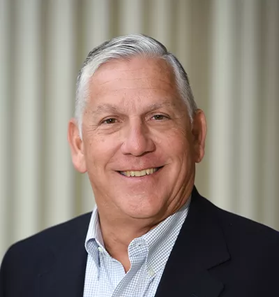 An older man with short gray hair smiles at the camera. He is wearing a dark blazer over a light-colored shirt. The background consists of vertical, light-colored blinds.