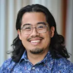 A person with long dark hair, glasses, and facial hair is smiling. They are wearing a blue shirt with a colorful pattern. The background shows vertical blinds in soft focus.