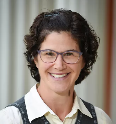 Person with short, curly dark hair, wearing glasses, a white shirt, and a dark vest, smiling against a blurred background.