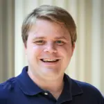 A smiling person with short, light brown hair wearing a navy blue polo shirt stands in front of a neutral background.