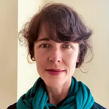 A person with short, wavy brown hair and a teal scarf stands in front of a neutral background.