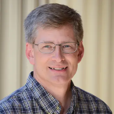 A man with short gray hair and glasses smiling at the camera. He is wearing a plaid shirt and standing in front of a light-colored curtain.