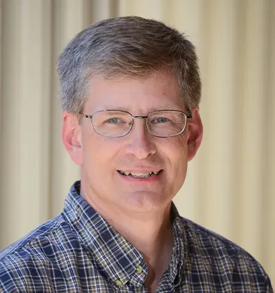 A man with short gray hair and glasses smiling at the camera. He is wearing a plaid shirt and standing in front of a light-colored curtain.