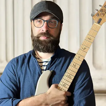 A bearded man wearing glasses and a cap holds a guitar, standing in front of a columned background. He's dressed in a blue shirt over a striped T-shirt.