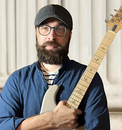 A bearded man wearing glasses and a cap holds a guitar, standing in front of a columned background. He's dressed in a blue shirt over a striped T-shirt.